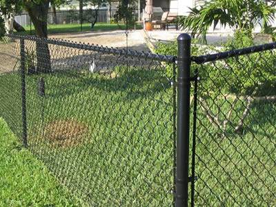 Black chain link fence with some thick posts is surrounding some trees and grass.